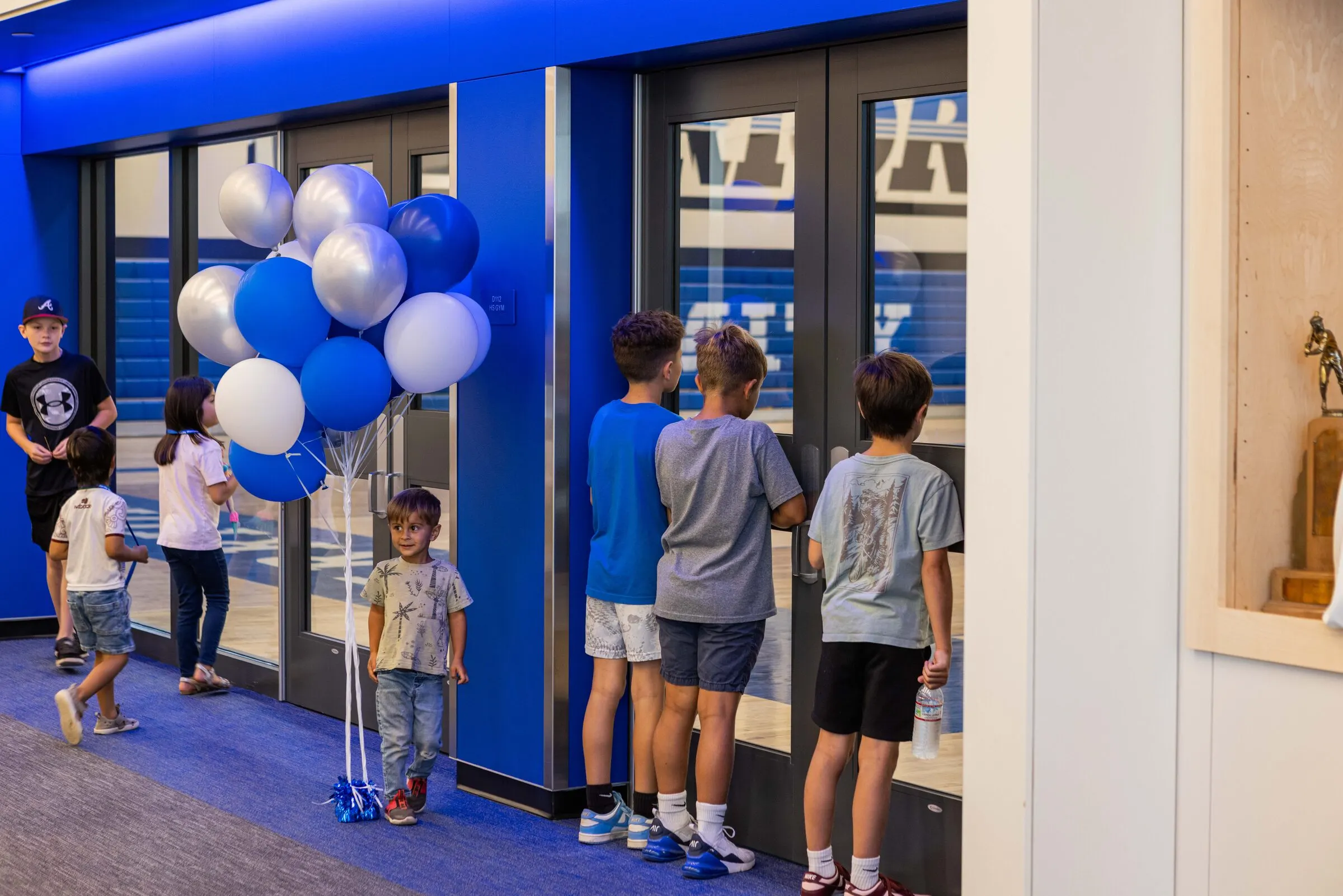 Young Amity students inside the new building on opening day — one boy holding royal-blue and white balloons, others looking eagerly through glass doors into the new gym, a trophy case visible at right.