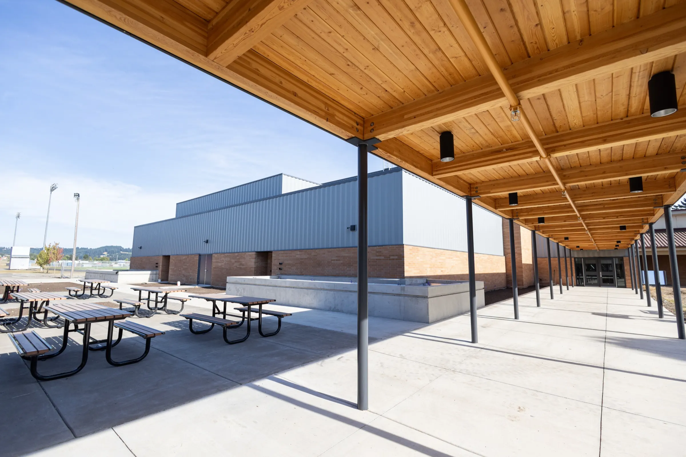 A covered walkway with a wood-plank ceiling runs along Amity High School, beside a courtyard of picnic tables and a new metal-sided building.