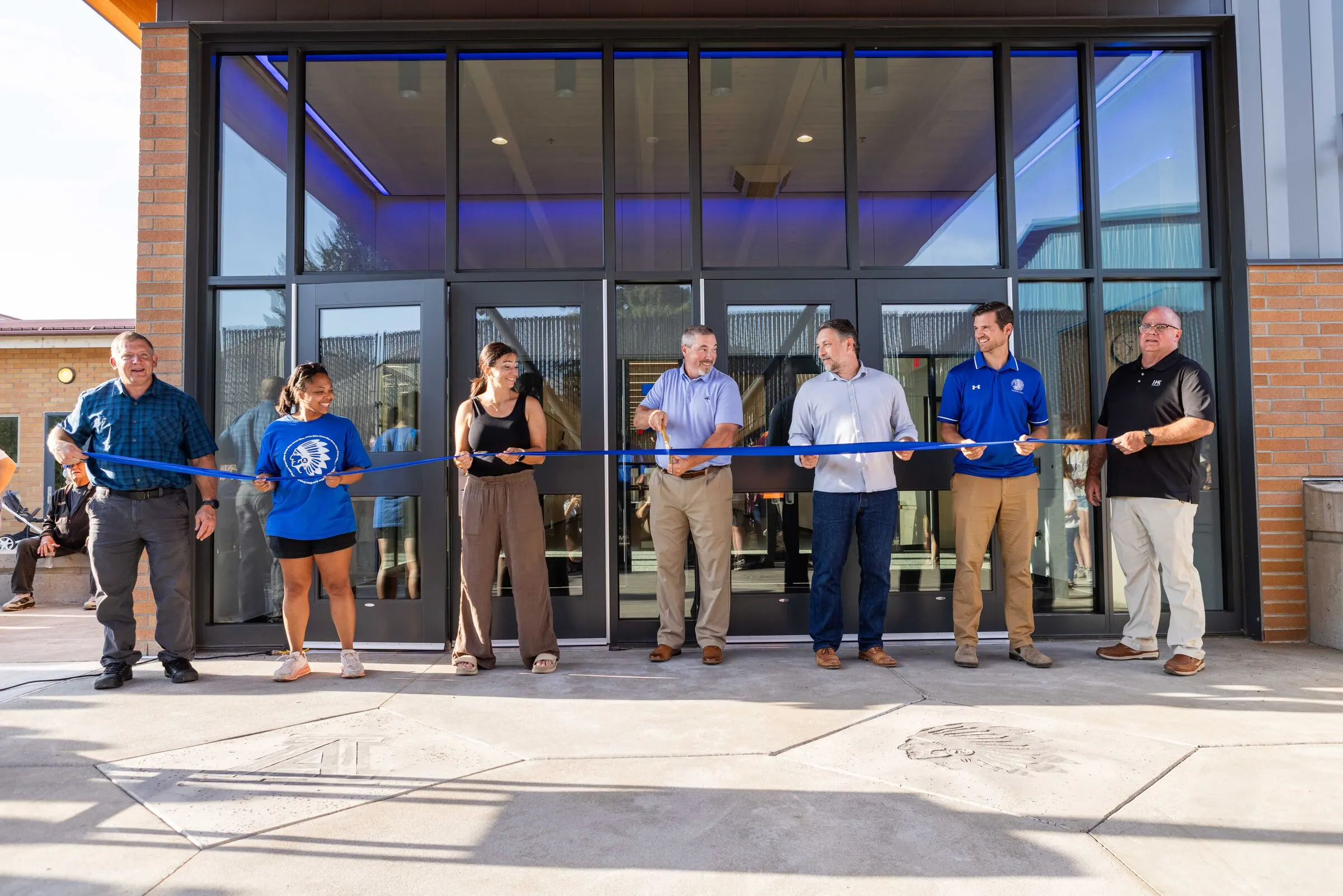 Seven community members stretching a blue ribbon across the front of the new glass entrance at Amity High School on opening day, the superintendent center-frame with scissors, the new lobby visible behind them.