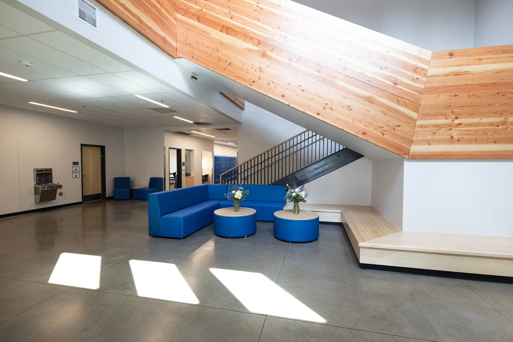Sunlit commons with blue seating beneath a wood-clad staircase.
