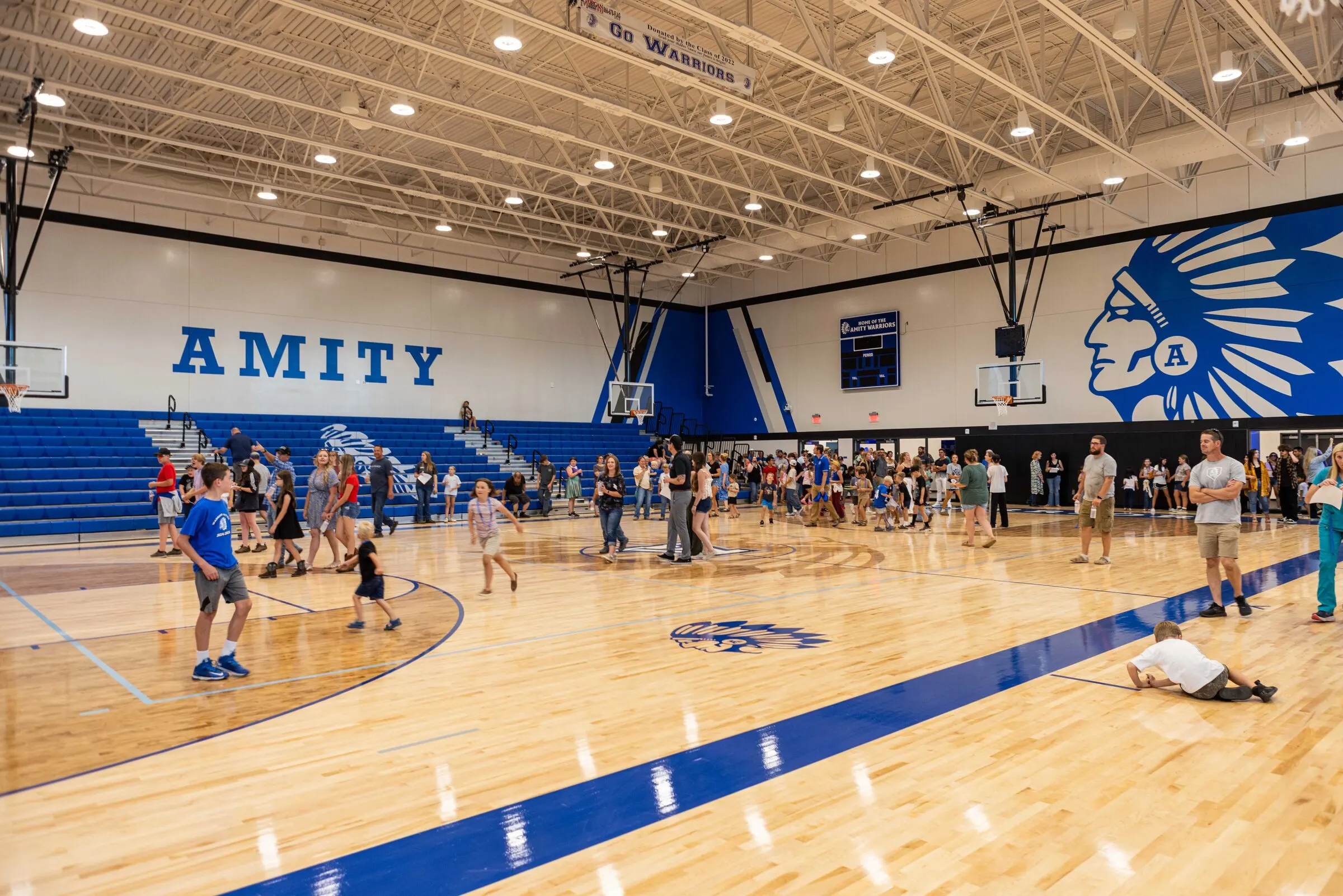 Inside the new Amity competition gym — the AMITY wordmark on the back wall, the Warrior headdress mural, families walking the polished maple court on opening day.